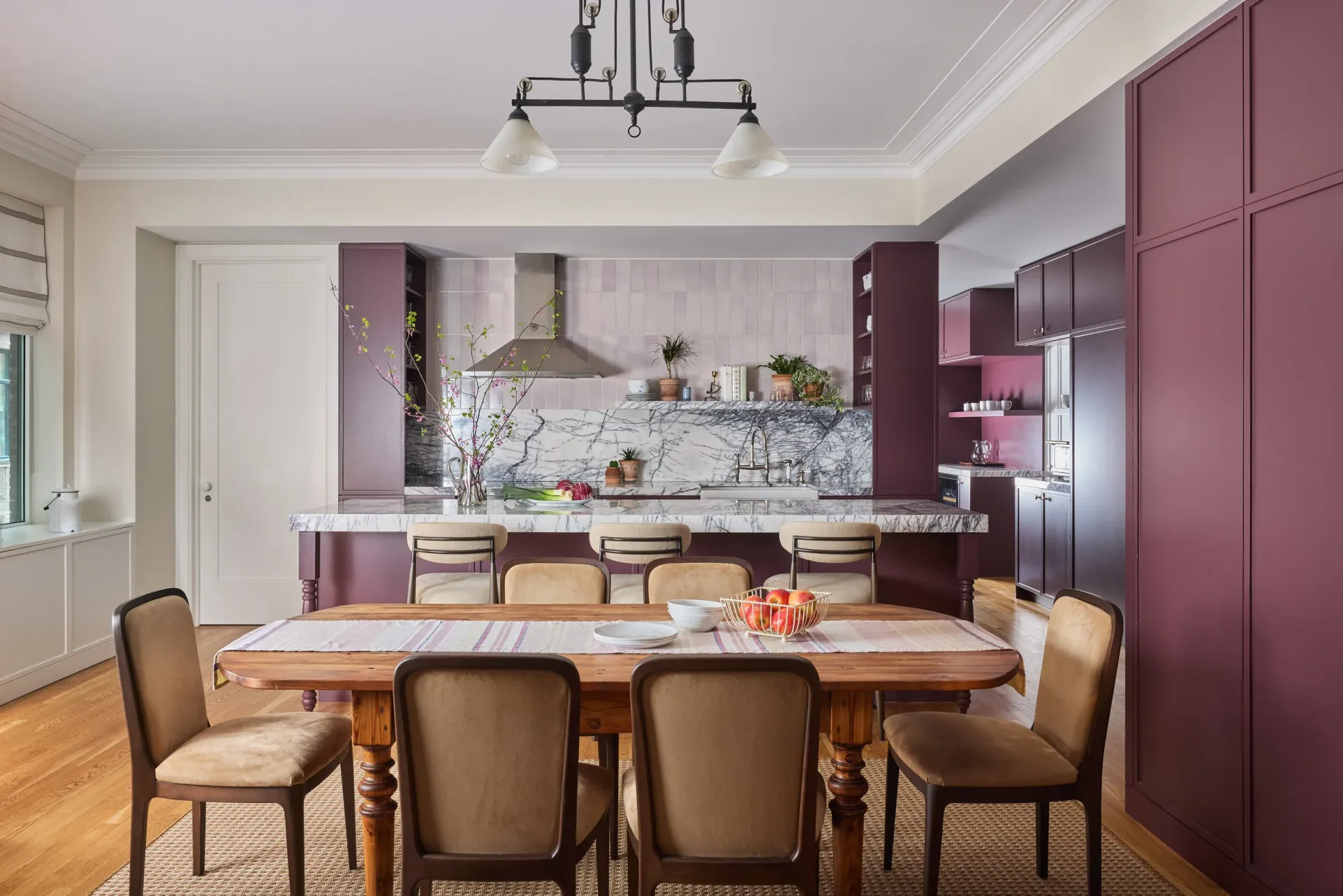 Modern kitchen and dining space featuring plum cabinetry, marble backsplash, wooden dining table with upholstered chairs, and pendant lighting by Liza Kuhn.