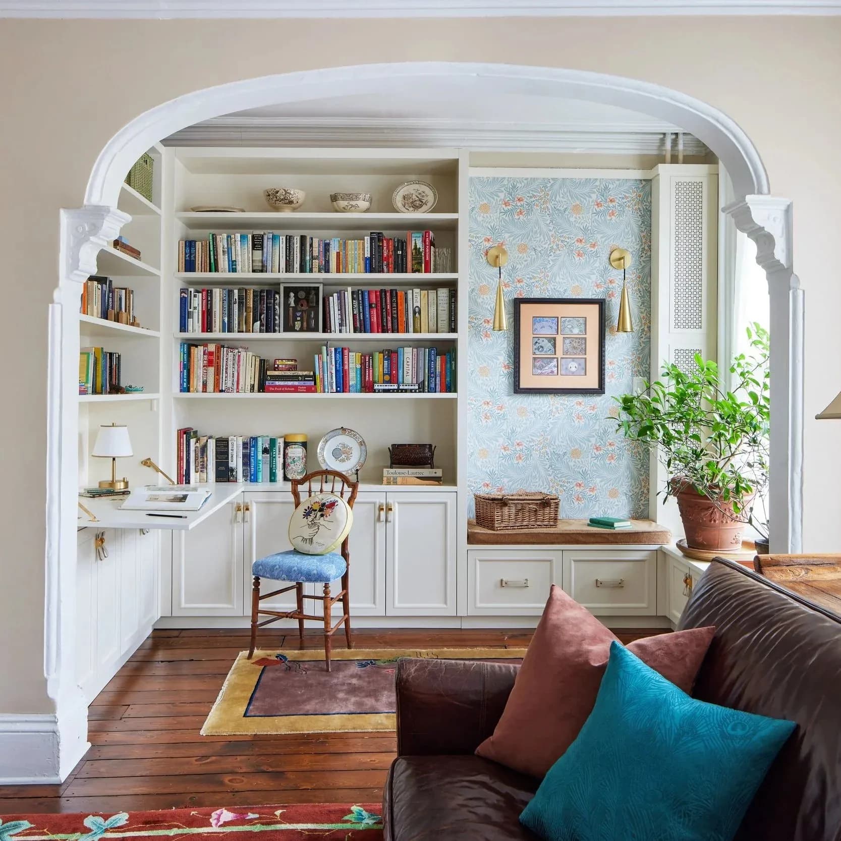 A bright alcove library with white built-in shelving filled with books, a blue floral wallpapered accent wall, potted plant, and desk area adjacent to a living room by Liza Kuhn.