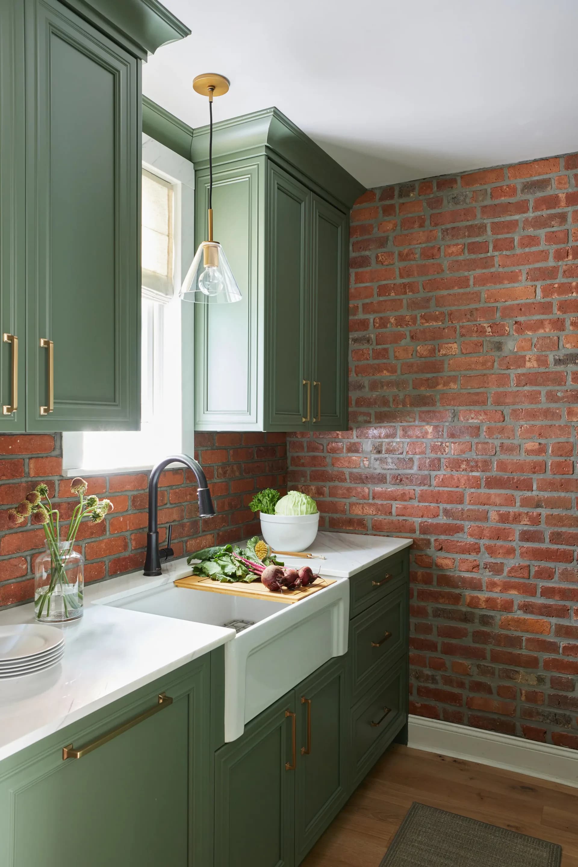 Modern kitchen with sage green cabinetry, white countertops, apron sink, and exposed red brick backsplash and accent wall, designed by Liza Kuhn.