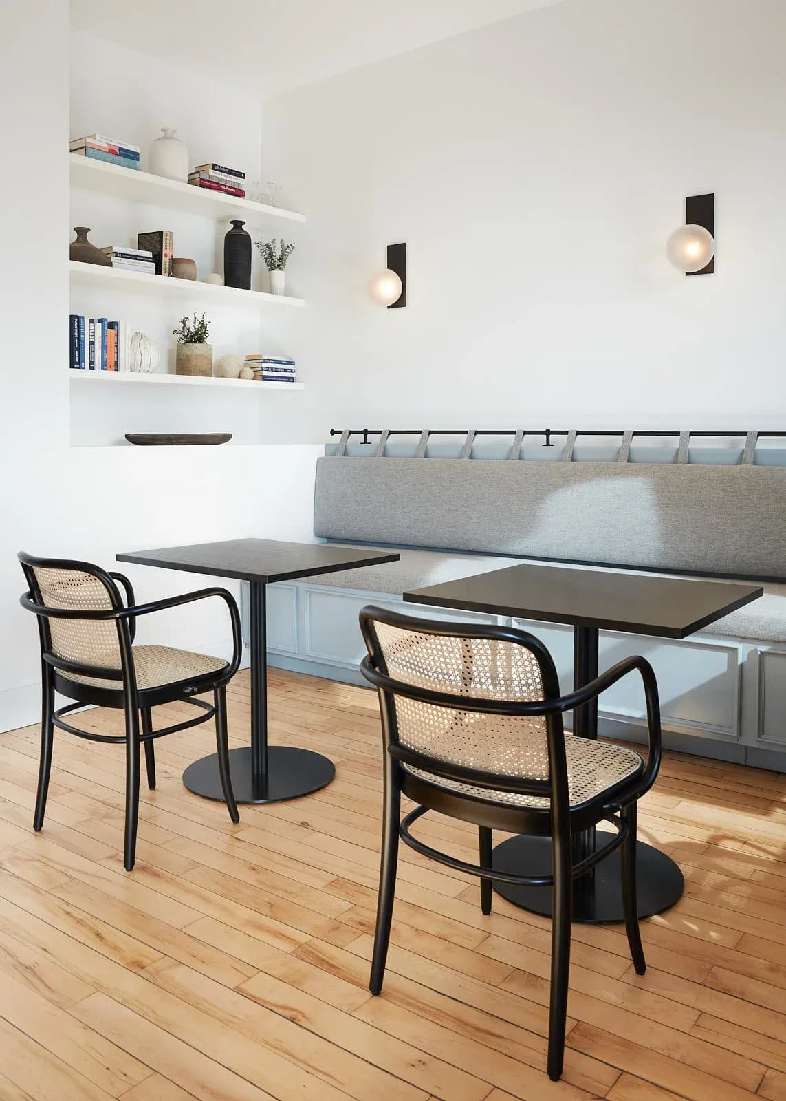 Modern café interior with gray upholstered banquette seating, black tables with rattan-back chairs, wooden flooring, and floating white shelving with décor by Lizi Gegenava.