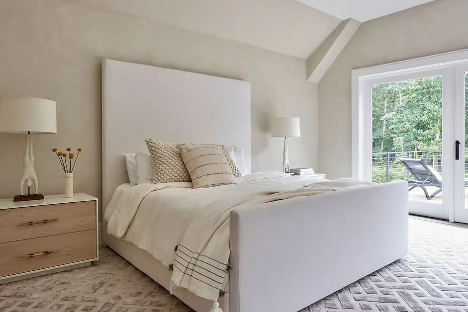 Serene master bedroom with white upholstered bed, neutral linens, natural wood nightstand, and glass doors opening to a balcony by Lo Chen.