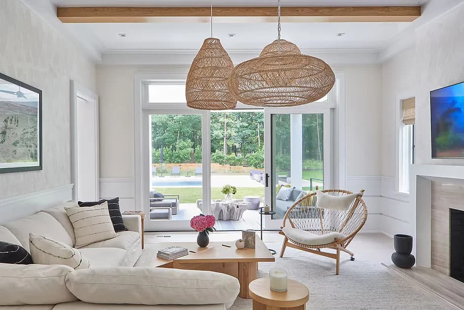 Bright modern living room with cream furnishings, woven pendant lights, wooden beams, and expansive glass doors overlooking green landscape, designed by Lo Chen.