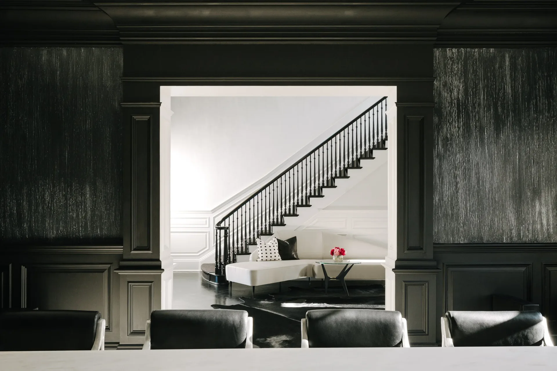 Modern staircase with black metal railing viewed through an open doorway, featuring white walls, minimalist furnishings, and a dark accent wall by Lori Margolis.