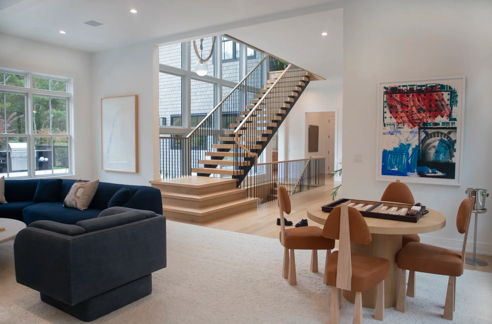 Modern two-story living space with navy sectional, wooden staircase, and dining area featuring natural wood furniture and contemporary artwork by Lori Margolis.