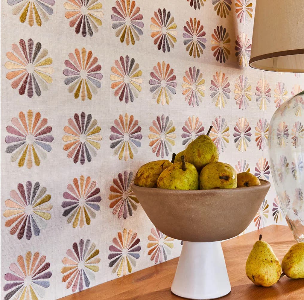 Warm-toned interior featuring patterned wallpaper with radiating fan motifs in earth tones, styled with a pedestal bowl of pears and scattered fruit, by Lori Weitzner.