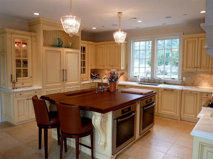 Cream-colored kitchen with large central island featuring dark wood countertop, stainless steel appliances, crystal pendant lighting, and cream cabinetry throughout, designed by Lorraine Frye.