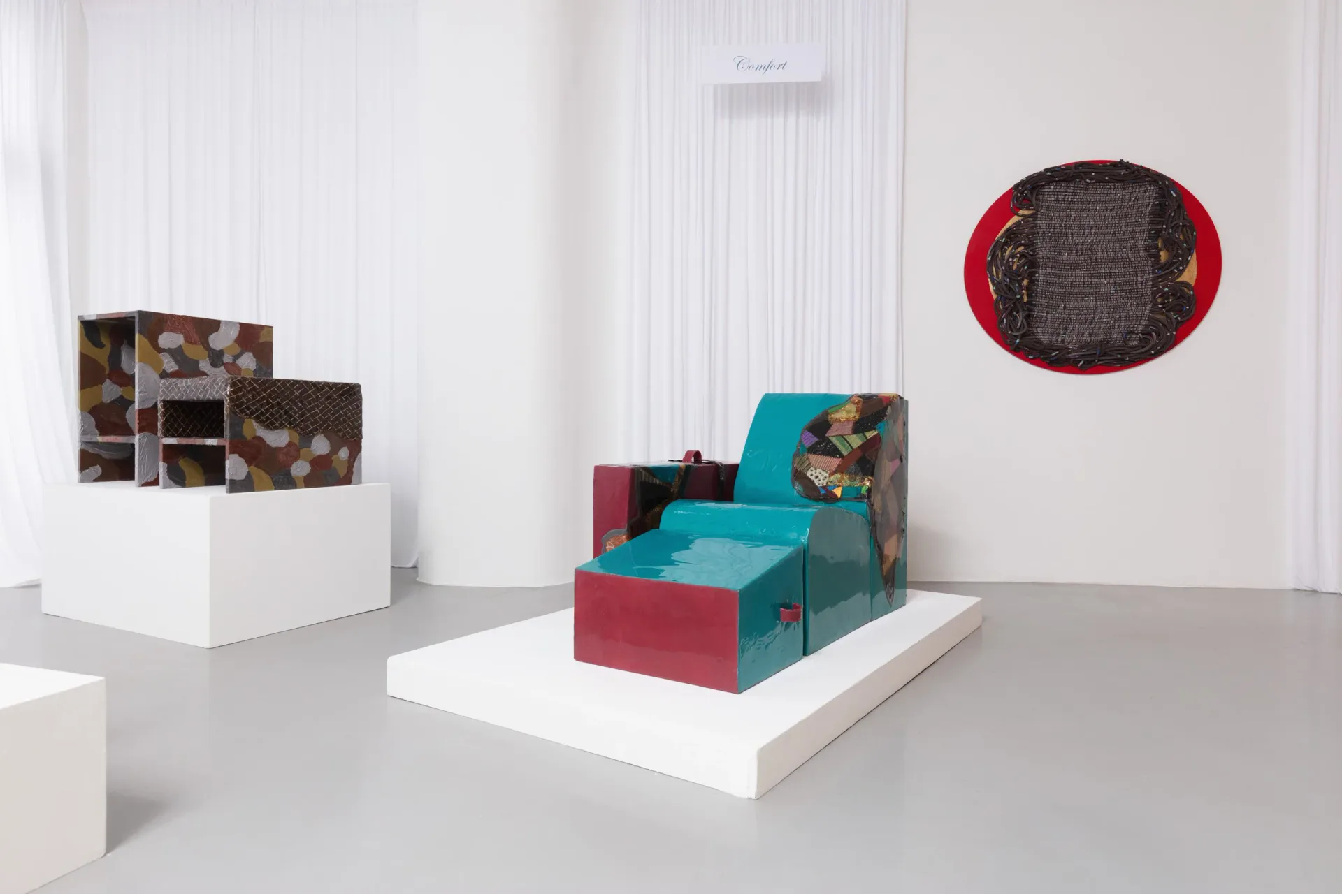 Gallery installation featuring modular furniture pieces in turquoise and burgundy with geometric forms on white platforms, alongside wall-mounted sculptural elements by Luam Melake.