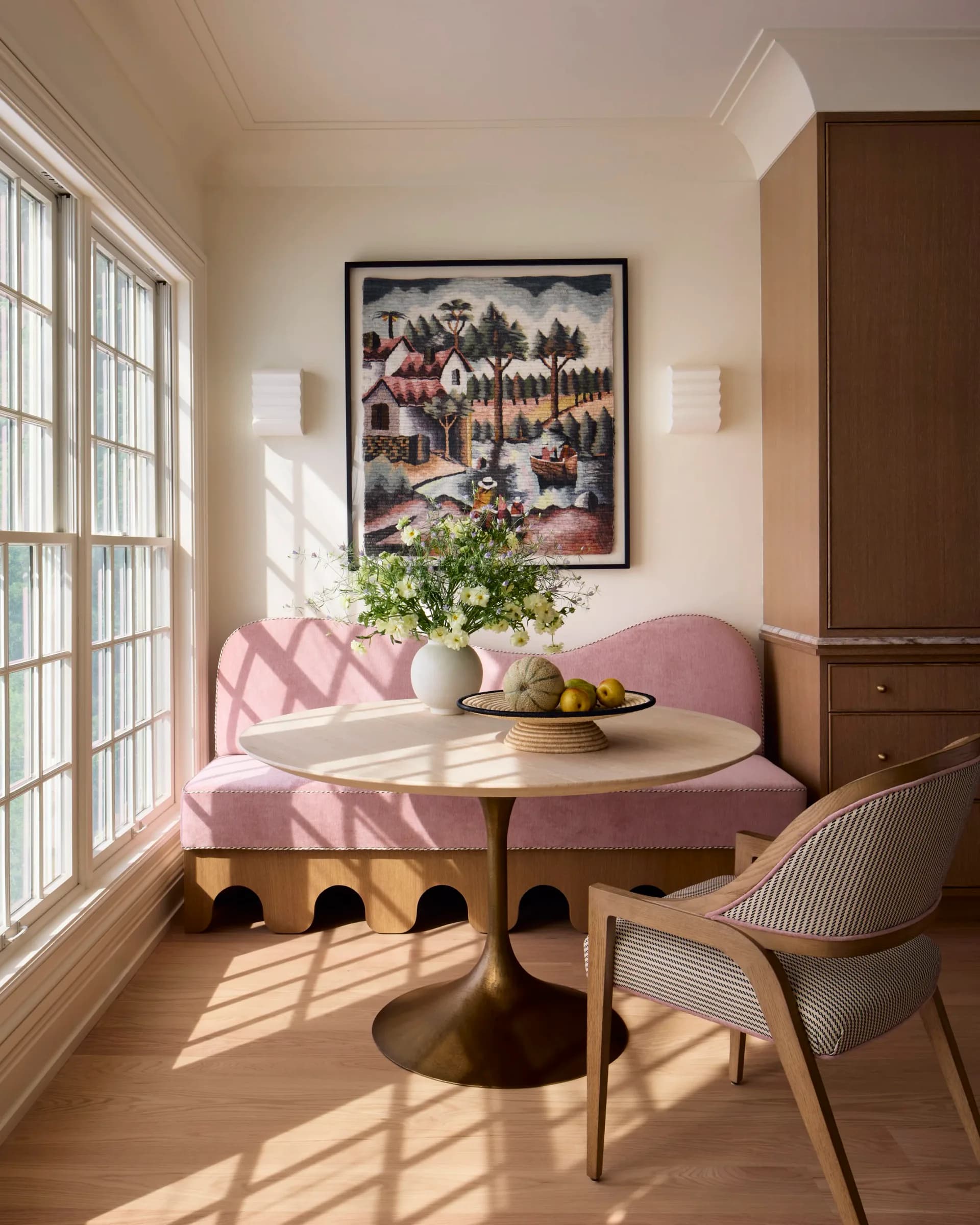 Bright dining nook with pink curved banquette, round marble table, natural wood chair, and large windows flooding the space with sunlight, designed by Luba Libarikian.