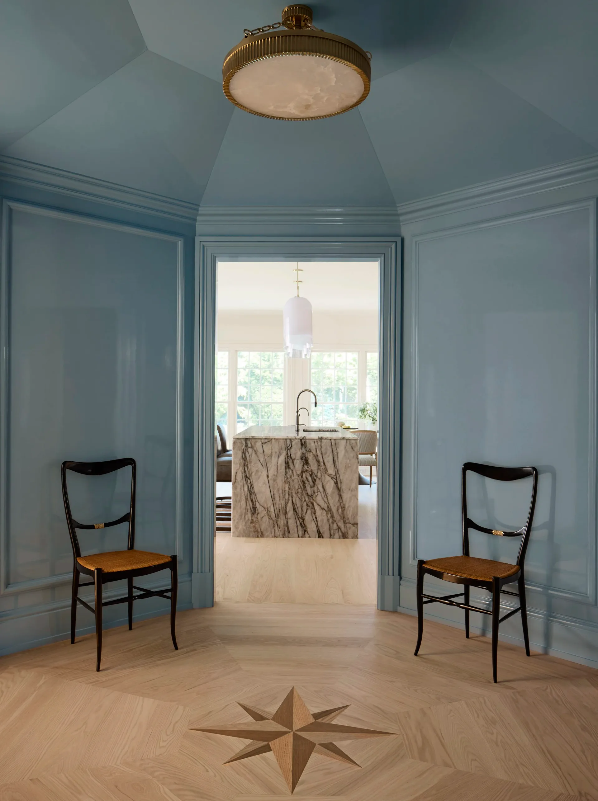 Light blue foyer with geometric inlaid wood floor, brass flush-mount ceiling fixture, and black chairs with woven seats flanking open double doors, designed by Luba Libarikian.