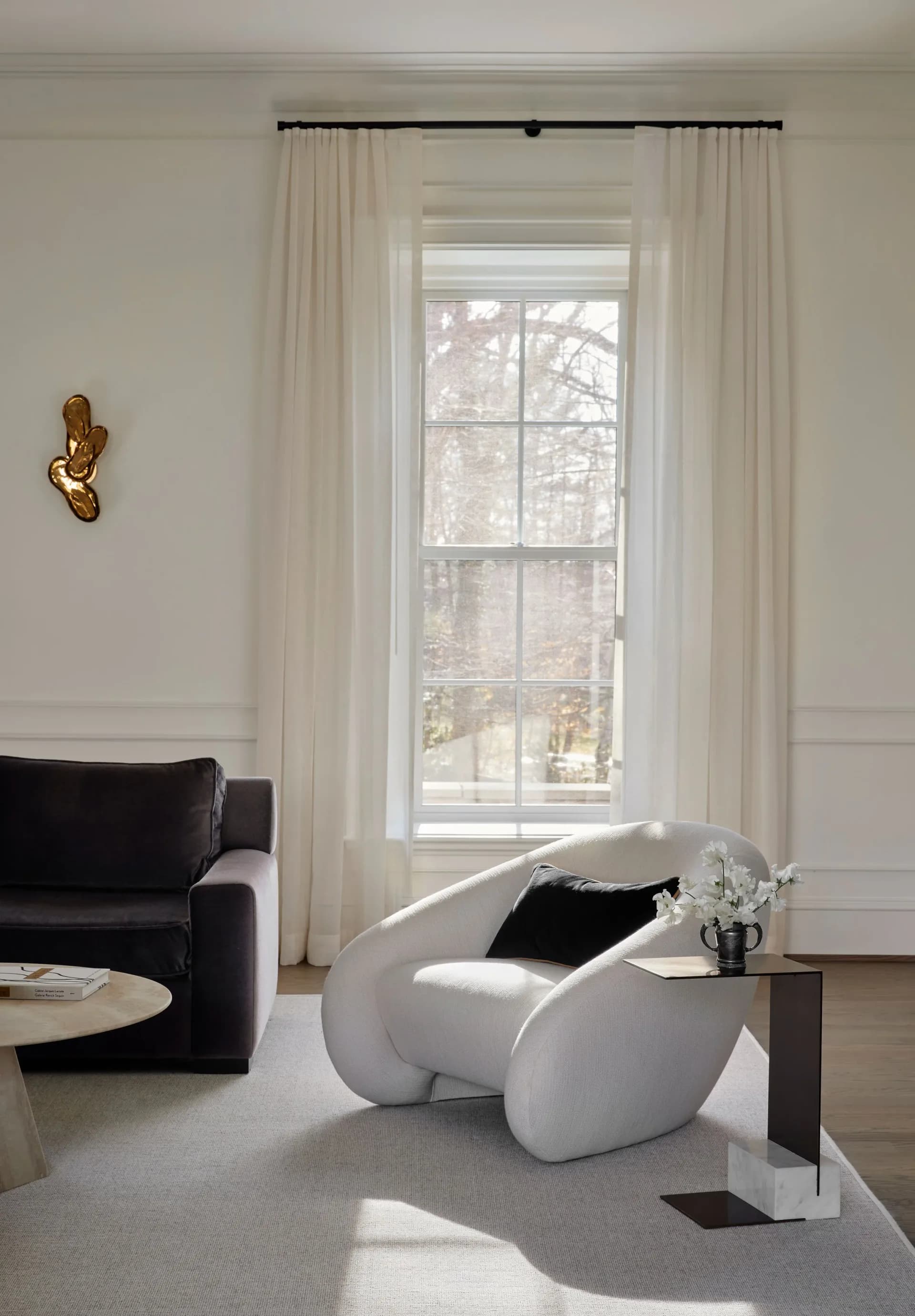 Minimalist living room with floor-to-ceiling windows, cream curtains, white curved armchair, black sofa, and sculptural decor by Luba Libarikian.