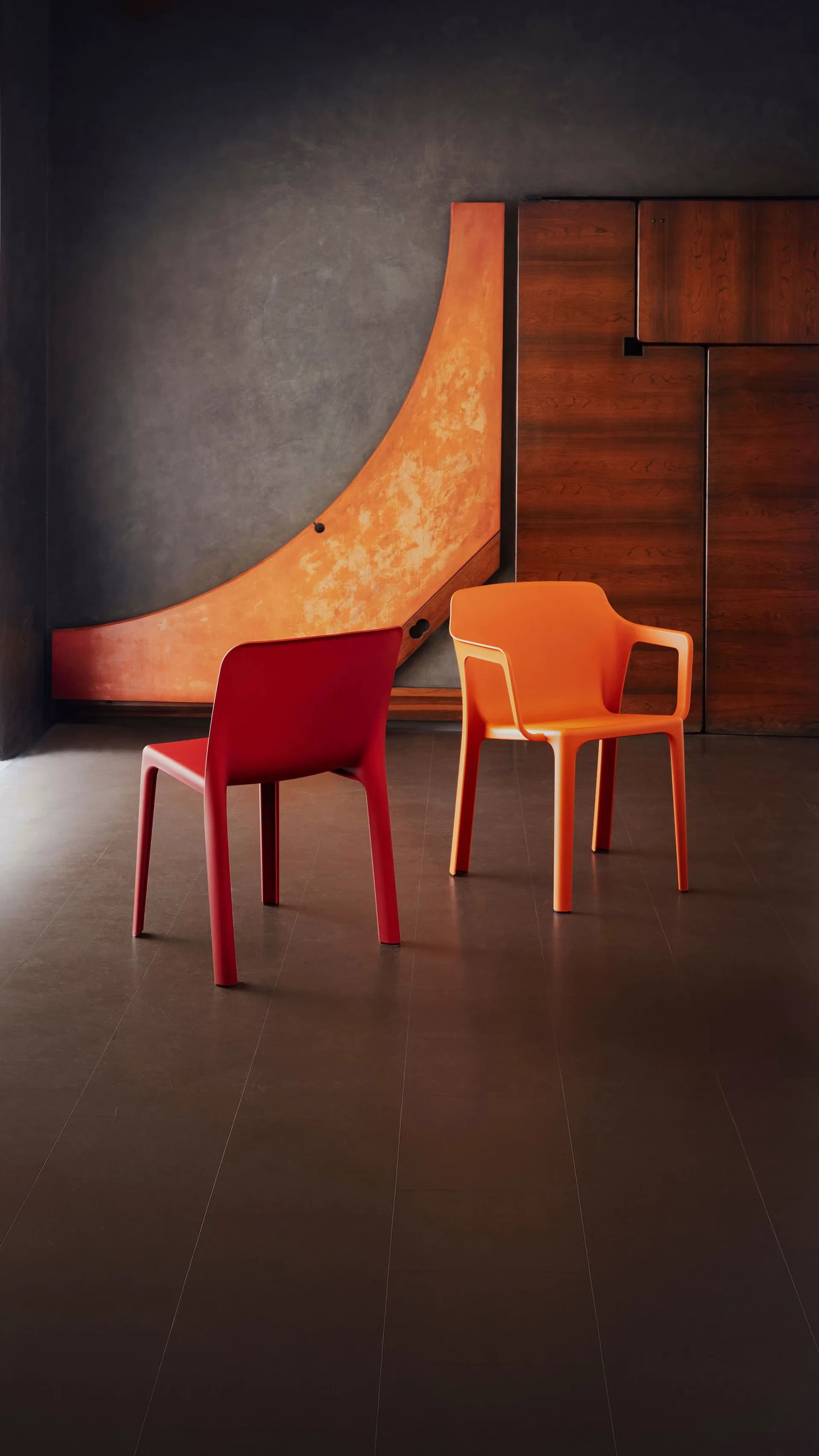 Modern interior with a red chair and orange chair positioned before a curved orange sculptural installation and wood-paneled wall by Luca Nichetto.