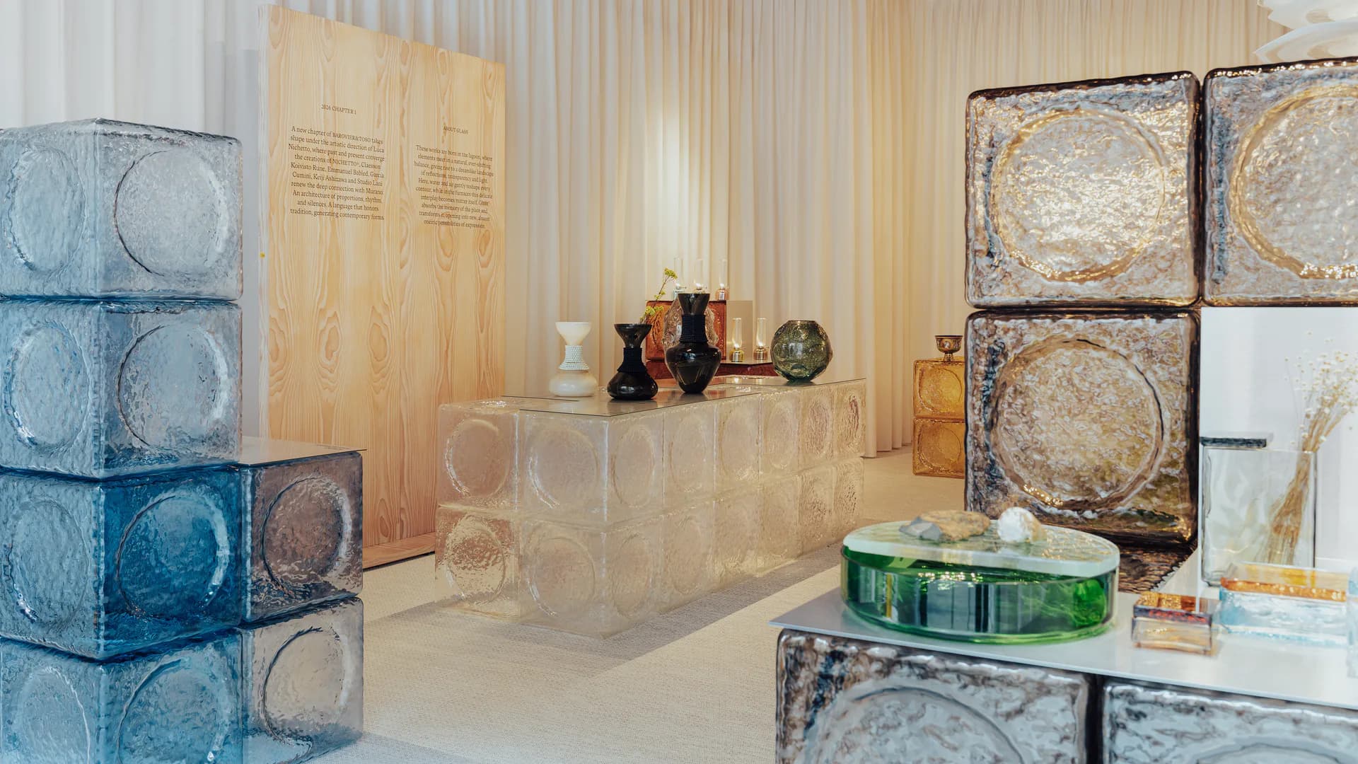 Contemporary exhibition space with stacked glass blocks, wooden panels, and circular map installations on white walls and floor by Luca Nichetto.