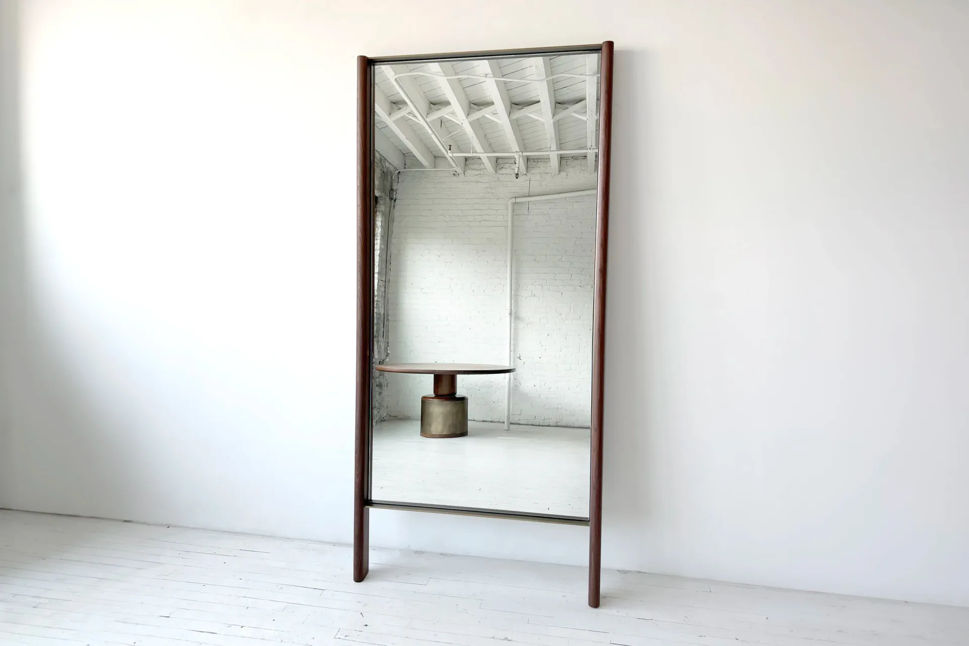 Minimalist wooden-framed mirror with metal fixture standing on white floor against white wall, a furniture design by Luke Fasano.