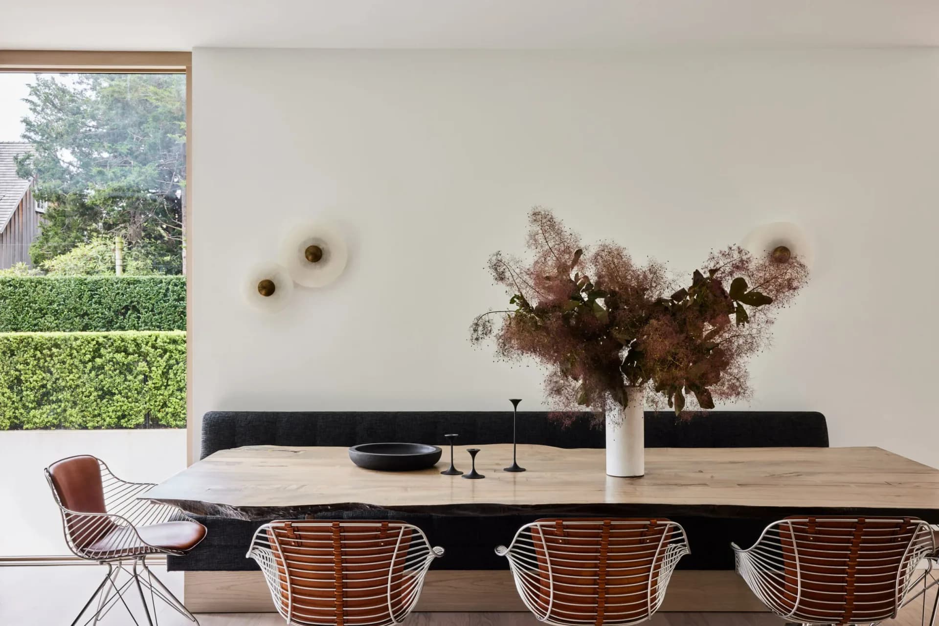 Minimalist dining space with natural wood table, woven rattan chairs, black upholstered bench, dried floral arrangement, and large window overlooking greenery by Luke Fasano.