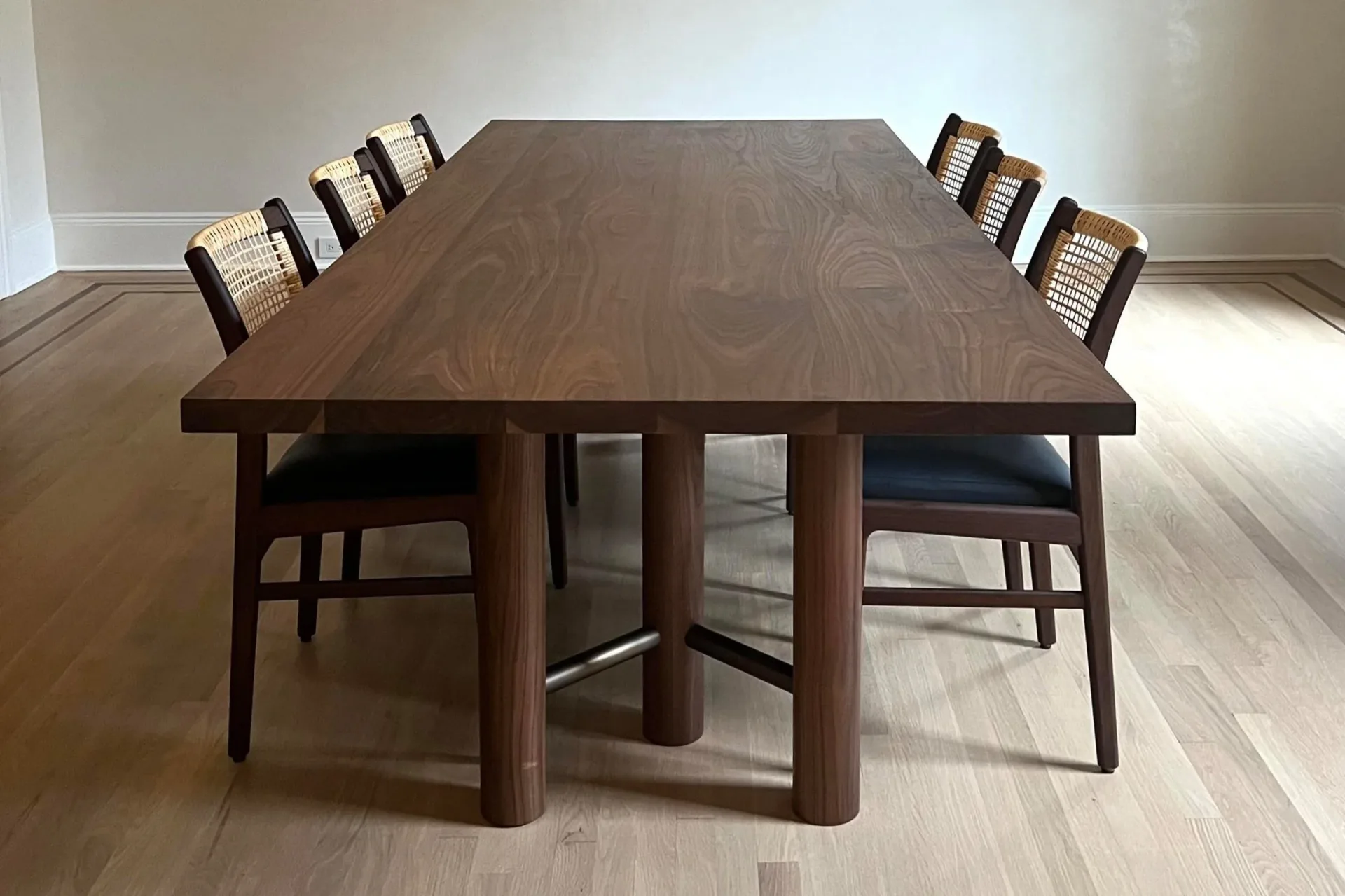 A minimalist dining room features a dark wood table with geometric base and matching chairs with woven backs, designed by Luke Fasano.