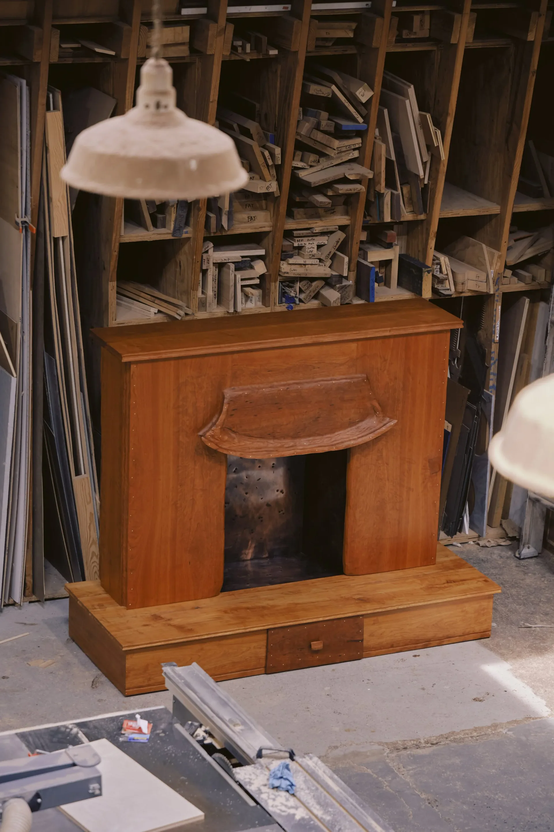 Wooden fireplace surround with integrated storage and bench base in warm honey tones, featuring a curved mantelpiece and arched opening, by Luke Malaney.