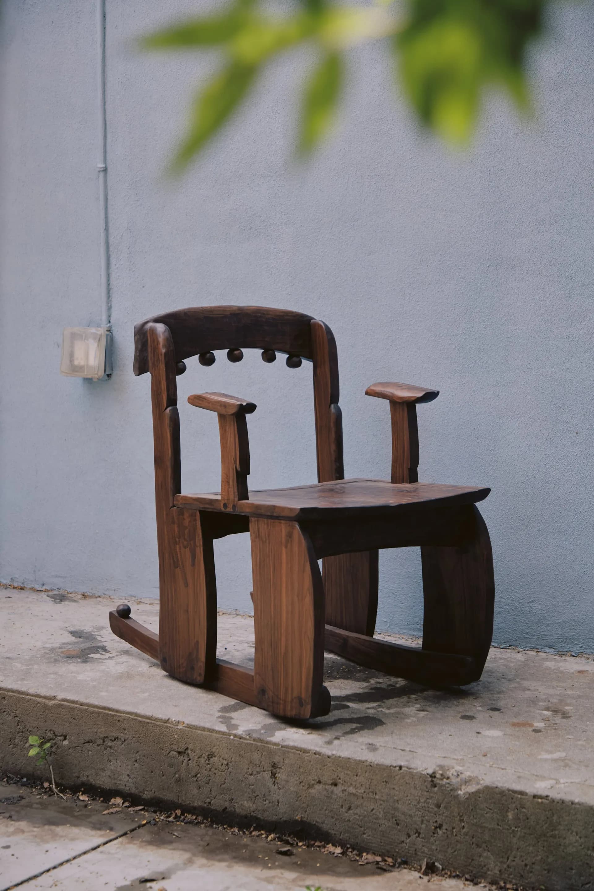 Dark wood sculptural chair with curved arms and geometric forms positioned against a white wall outdoors by Luke Malaney.