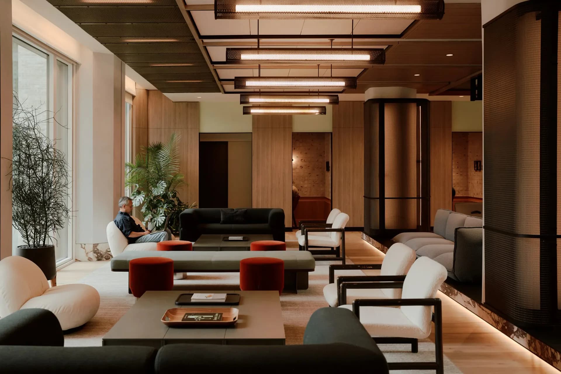 Warm-toned modern lounge with wood paneling, linear ceiling lights, and contemporary furniture in cream and burgundy by Khanh Luu.