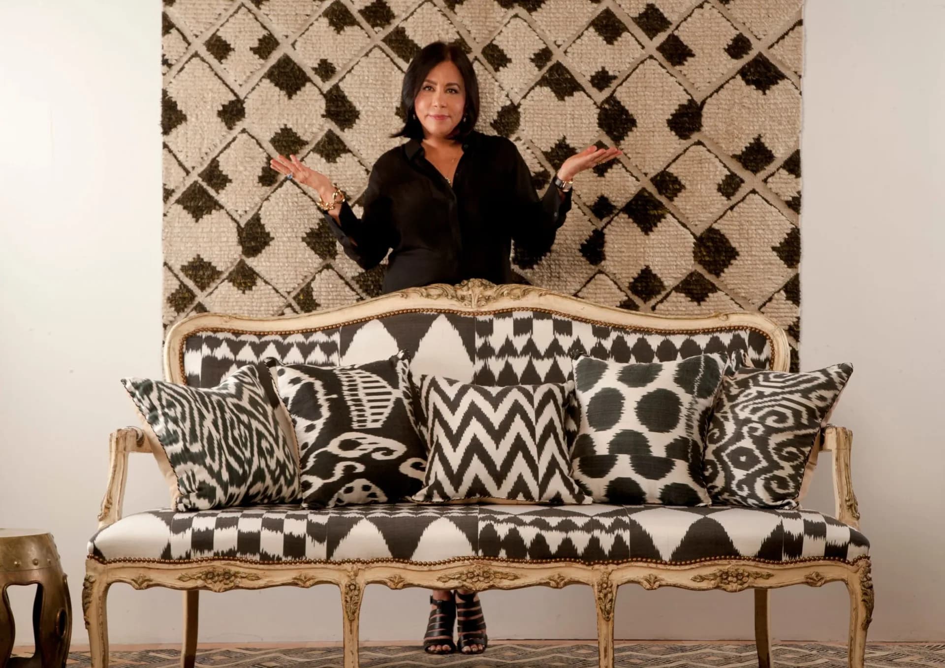 Black and white patterned pillows displayed on a natural wood frame sofa against a geometric wallpapered backdrop, by Madeline Weinrib.