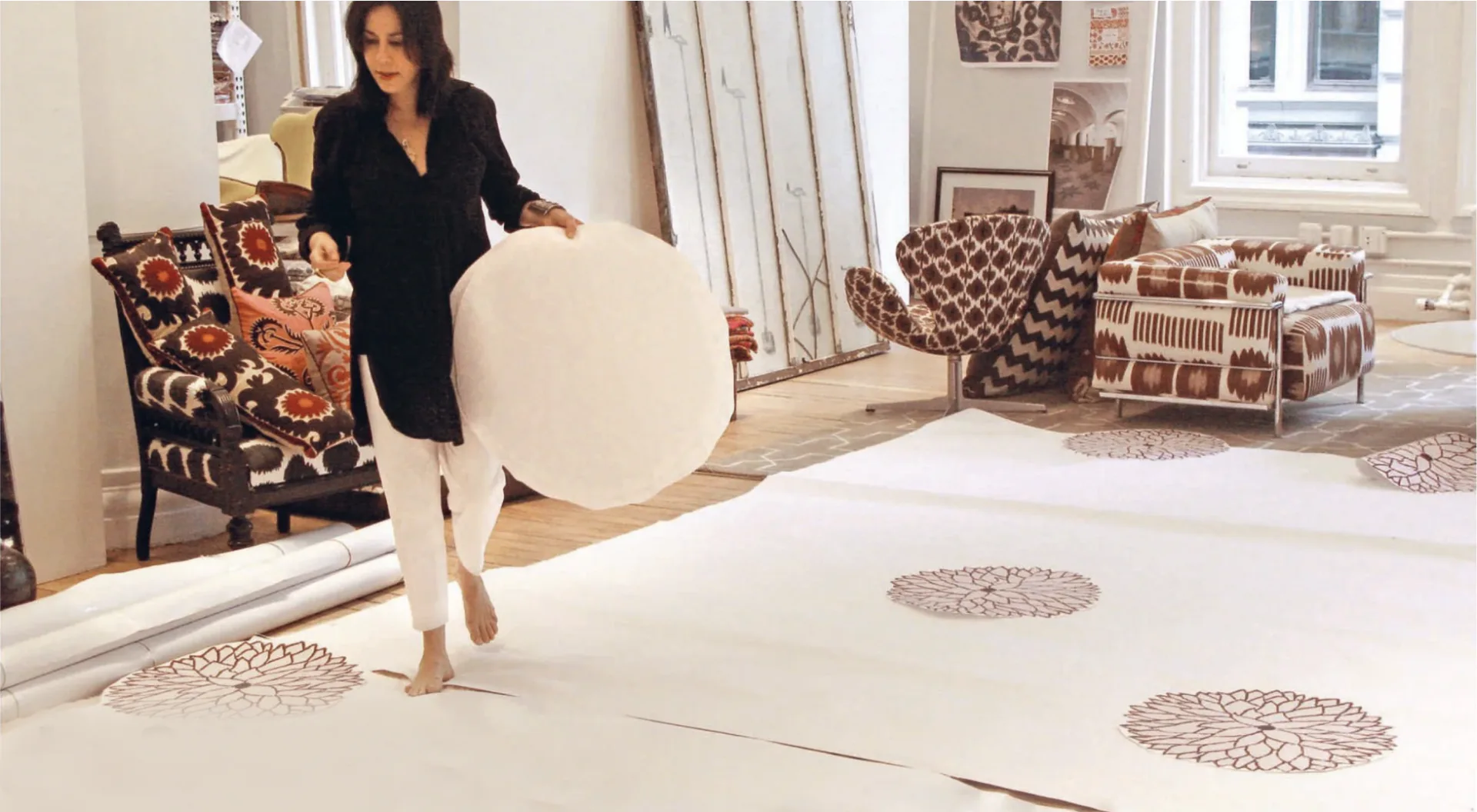 Designer arranges patterned textile samples on a white floor in a bright studio space with furnishings by Madeline Weinrib.