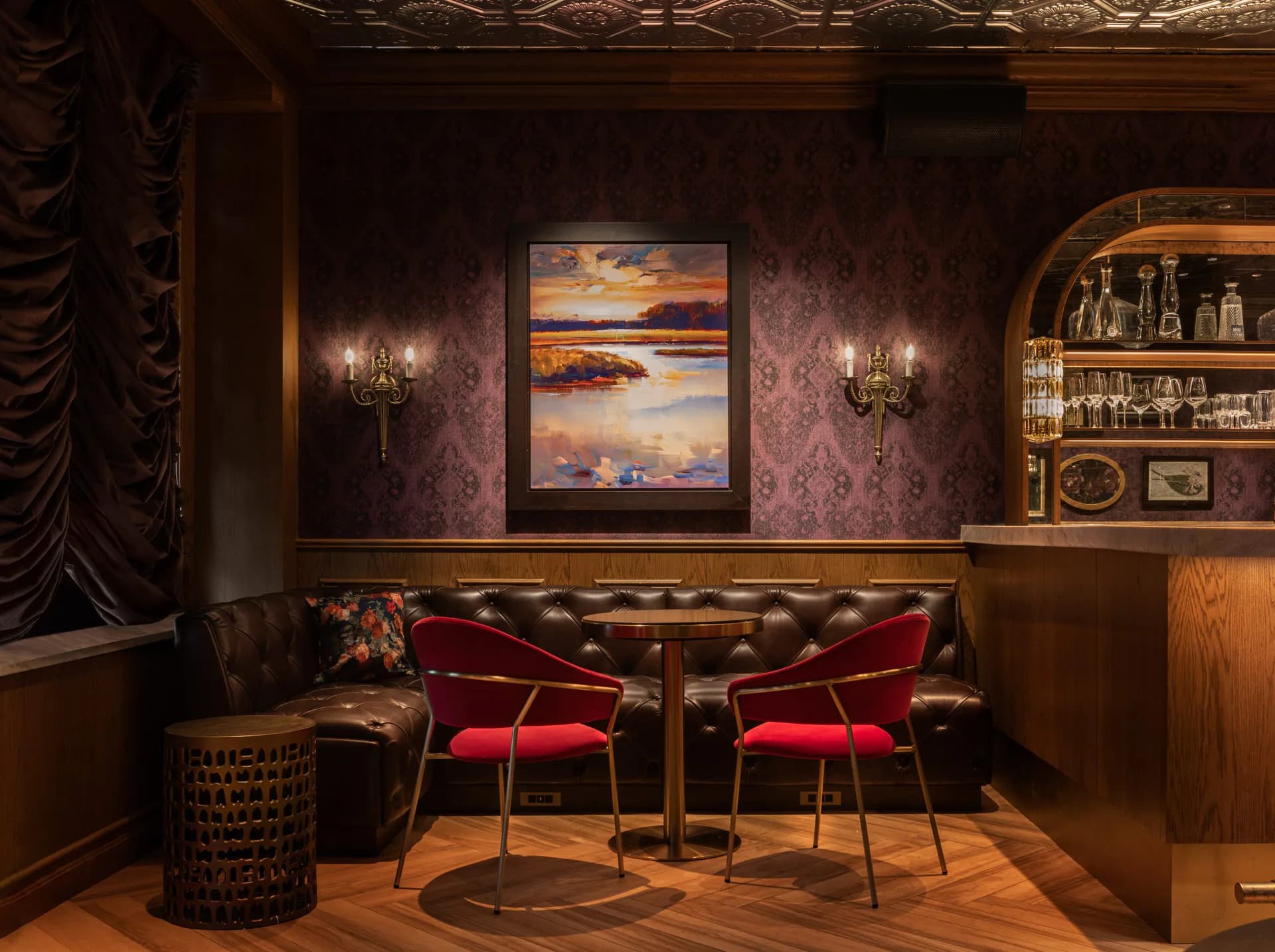 Luxurious lounge interior with burgundy velvet chairs, leather sofa, brass sconces, ornate ceiling detail, and framed sunset artwork by Mahek Joshi.