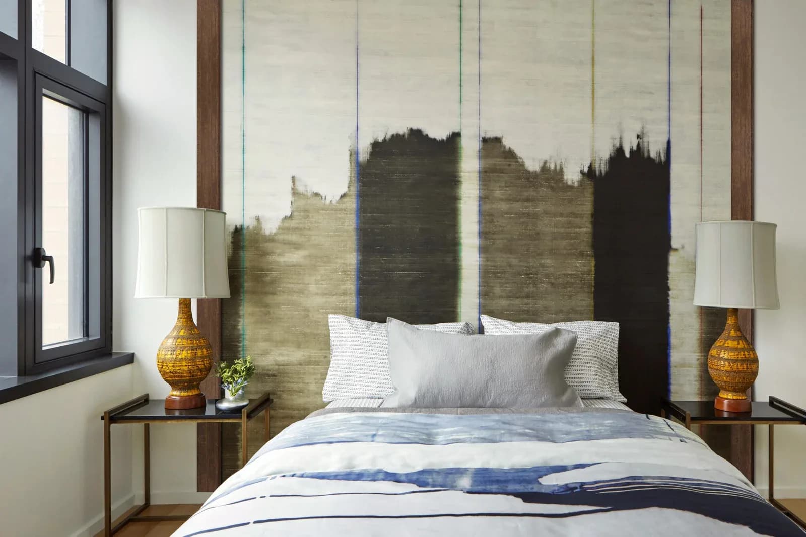 Modern bedroom with custom landscape headboard artwork, pendant lighting, brass table lamps, and blue bedding by Mahnoor Jahangir.