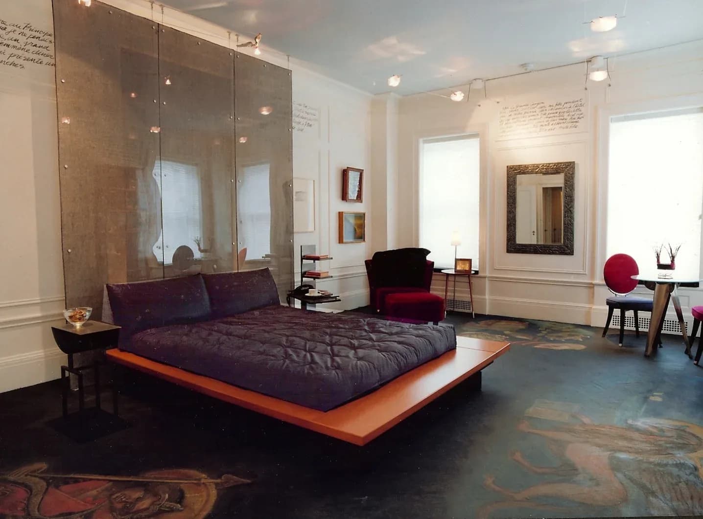 Modern bedroom with black quilted platform bed, orange base, glass partition headboard, burgundy accents, and teal floor installation by Marc Donnenfeld.