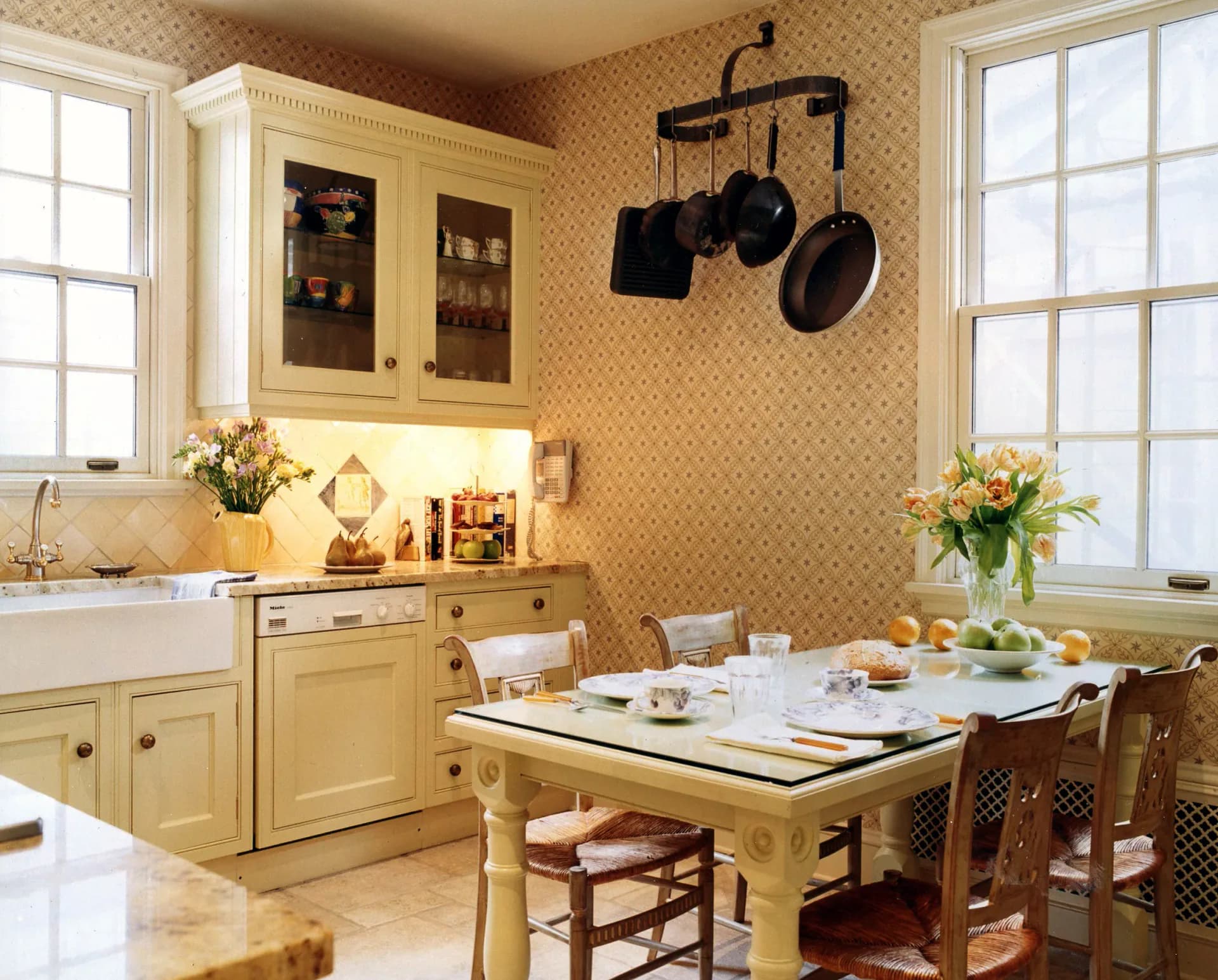 Warm yellow kitchen with patterned wallpaper, cream cabinetry, dining table with place settings, and pendant pot rack, designed by Marc Donnenfeld.