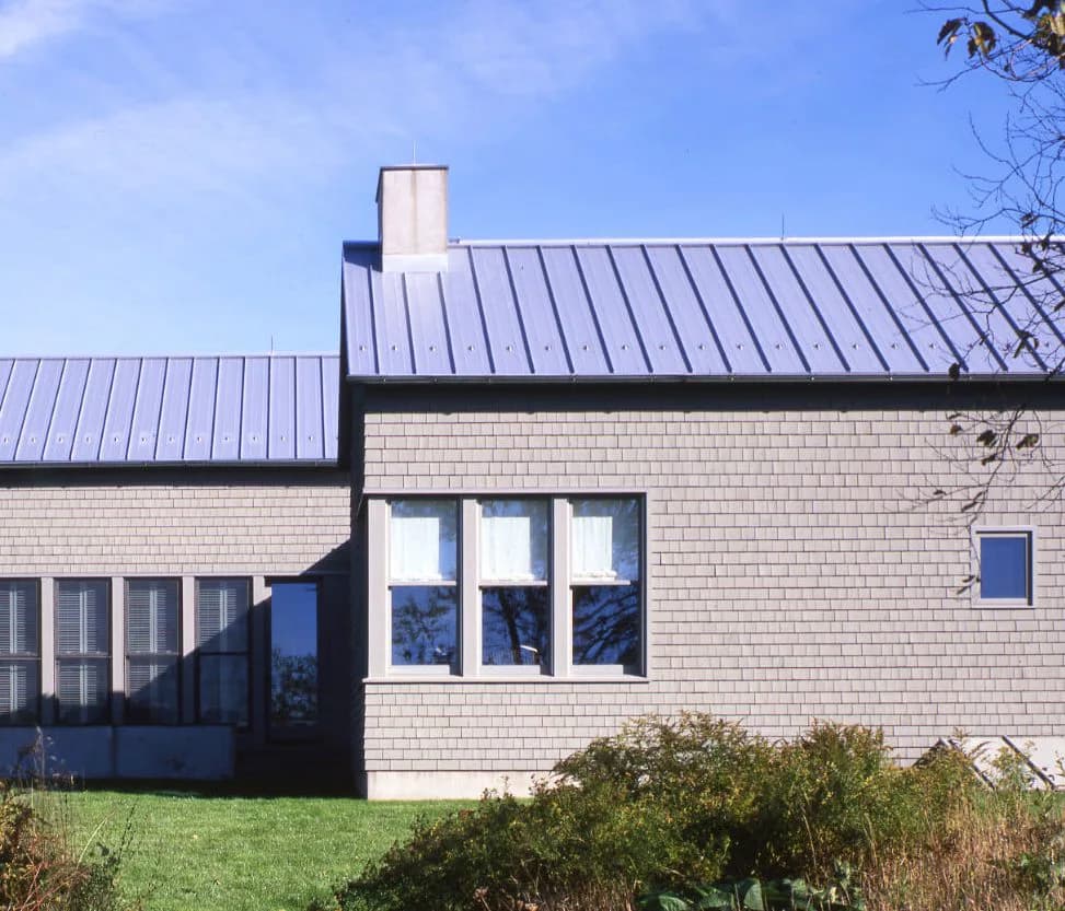 Modern residential architecture with cream brick facade, standing seam metal roof, and large window wall overlooking landscaped grounds by Marc Leff.
