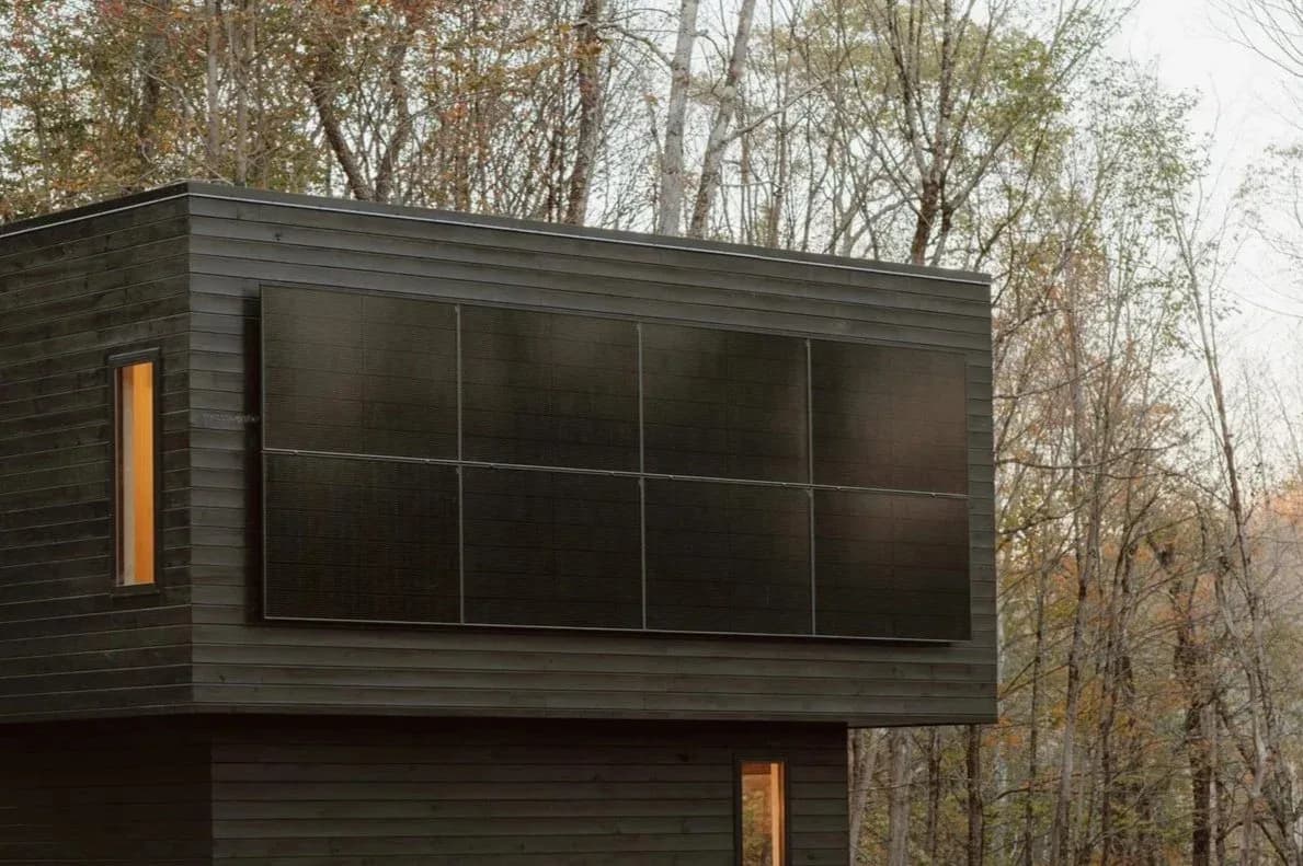 Modern dark-clad residential structure with expansive gridded black windows and warm wood accents nestled among bare trees by Marc Thorpe.