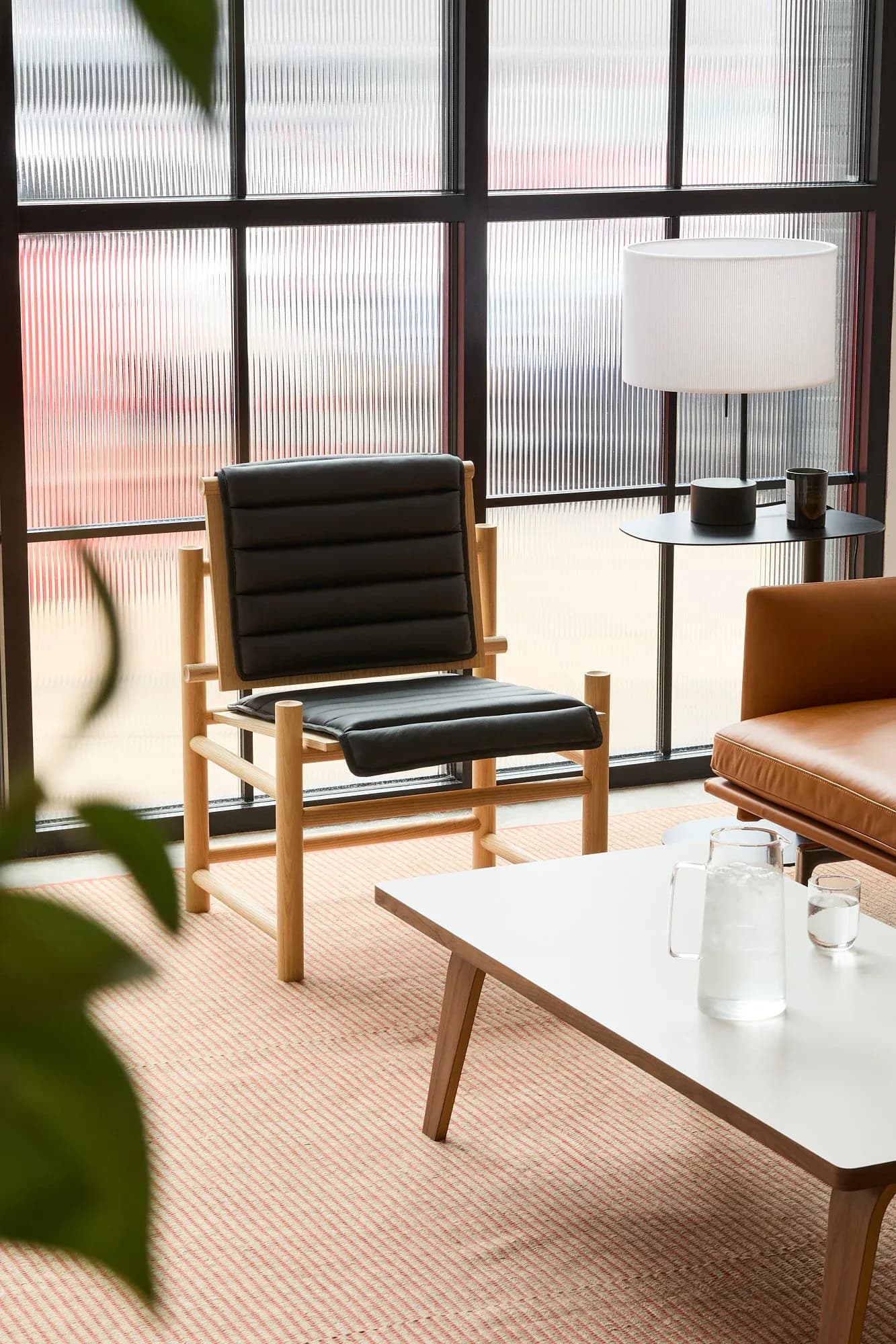 Minimalist interior with natural wood-framed lounge chair, black leather seat, white side table, and floor lamp beside large windows with translucent shading by Maren Jevremov.