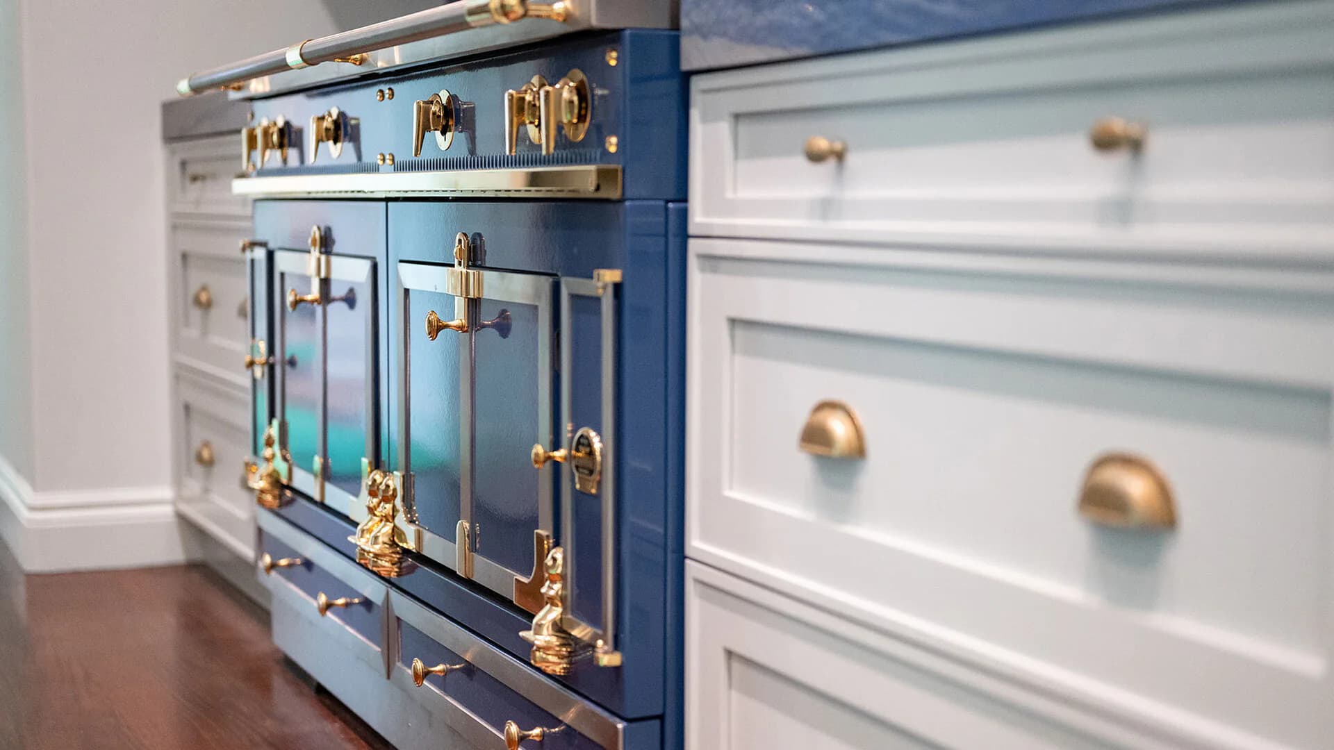Modern kitchen cabinetry featuring a blue-painted range hood with brass hardware and accents, paired with white shaker-style cabinetry with brass pulls by Margaret Costello.
