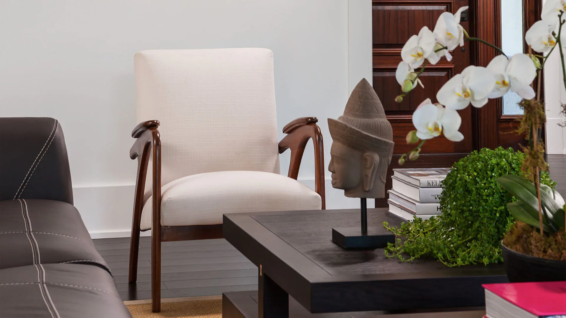 Modern living room with cream upholstered armchair, dark wood frame, black side table, white orchids, decorative objects, and neutral palette by Margaret Costello.