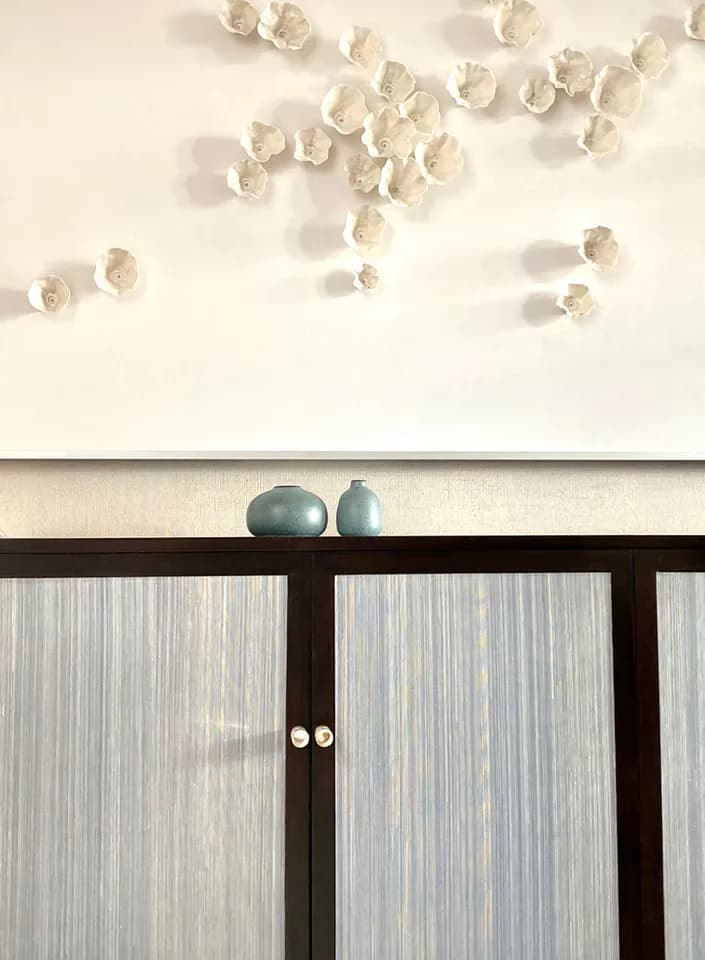 Minimalist interior with cream wall featuring sculptural ceramic shell installations above a dark wood cabinet with frosted glass doors and two blue ceramic vessels, by Margarita Rael.