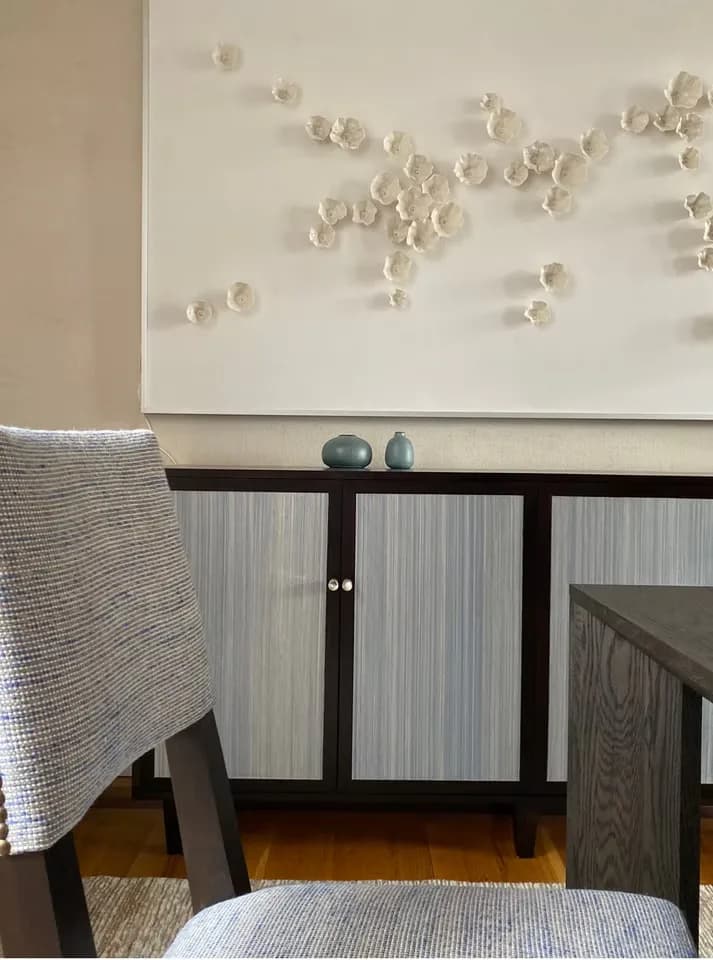 Contemporary interior featuring a black credenza with ribbed glass doors, cream textured seating, and an abstract wall installation of white sculptural forms by Margarita Rael.