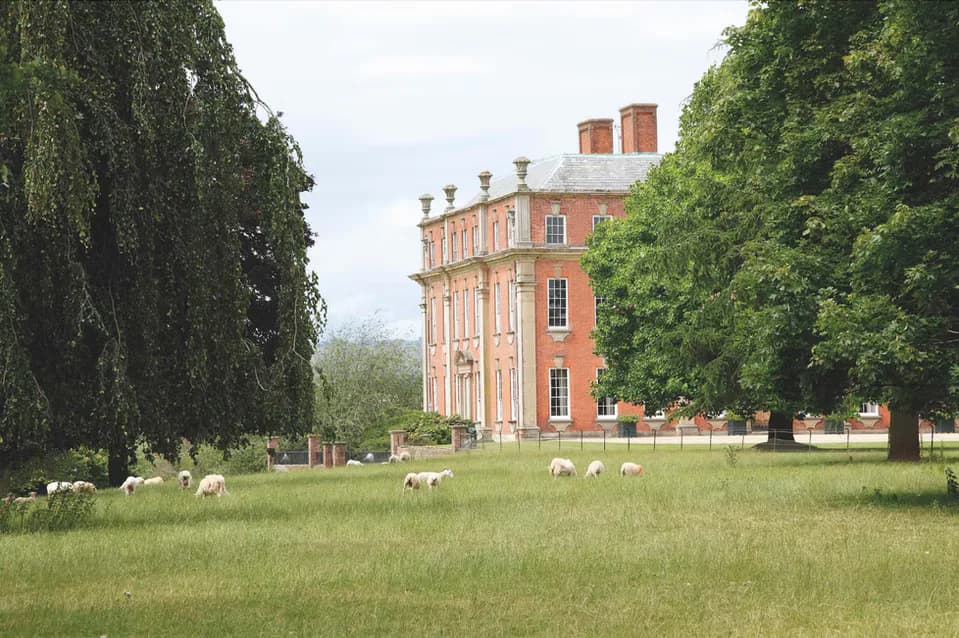 Neoclassical brick estate with symmetrical facades overlooks manicured lawn dotted with grazing sheep, framed by mature trees by Maria Filar.