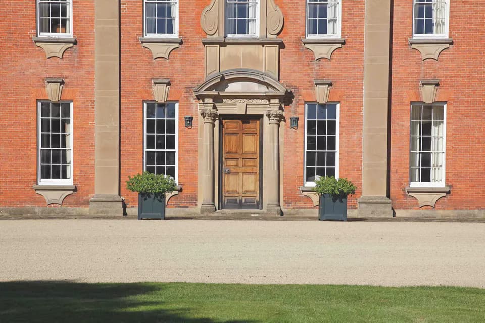 Symmetrical red brick Georgian facade with sash windows, central arched doorway, flanking planters, and manicured lawn by Maria Filar.