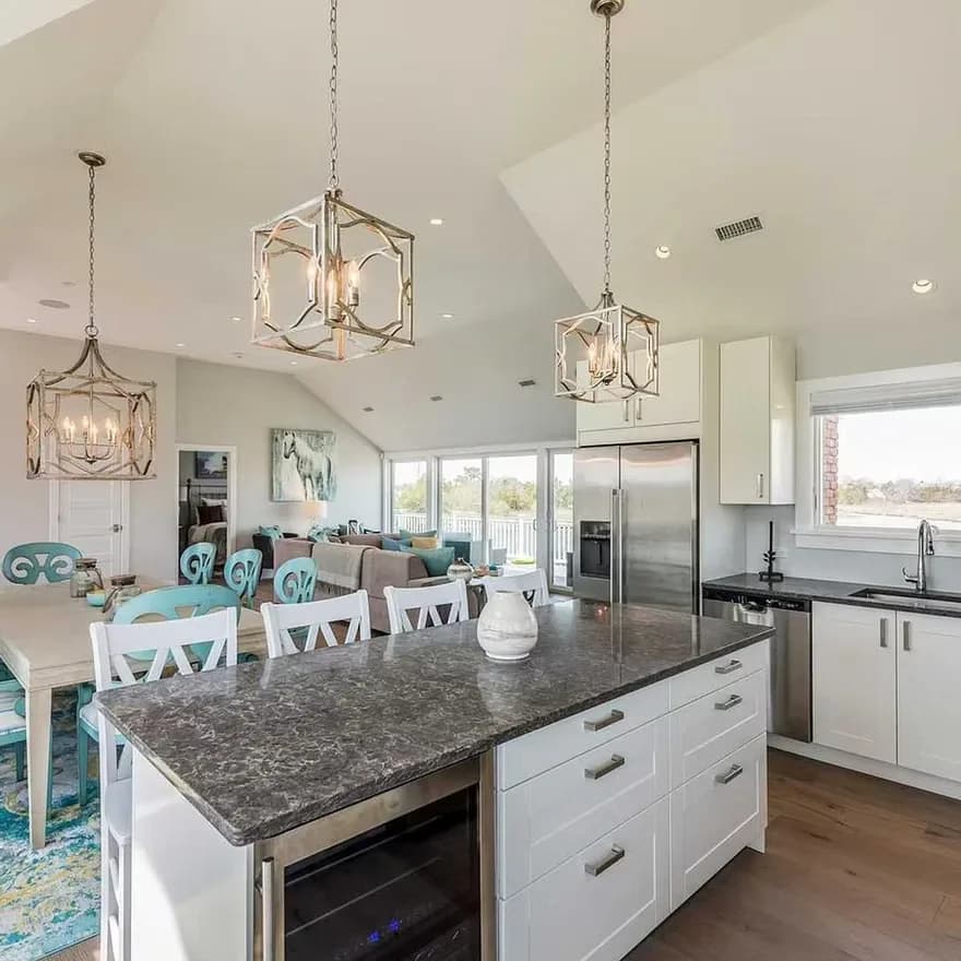 Open-concept kitchen and dining area with white cabinetry, gray granite island, geometric pendant lights, turquoise dining chairs, and expansive windows overlooking landscape by Mariama Agi.