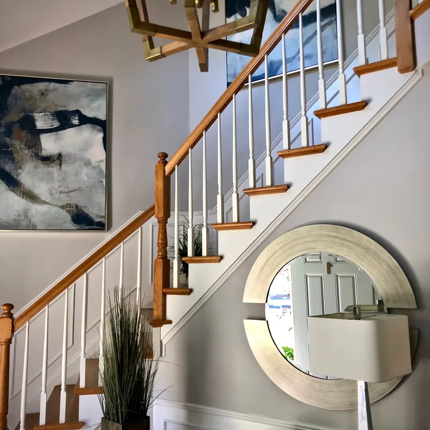 Modern staircase with wooden handrail and metal balusters, white walls, round mirror, abstract artwork, and pendant lighting by Mariama Agi.