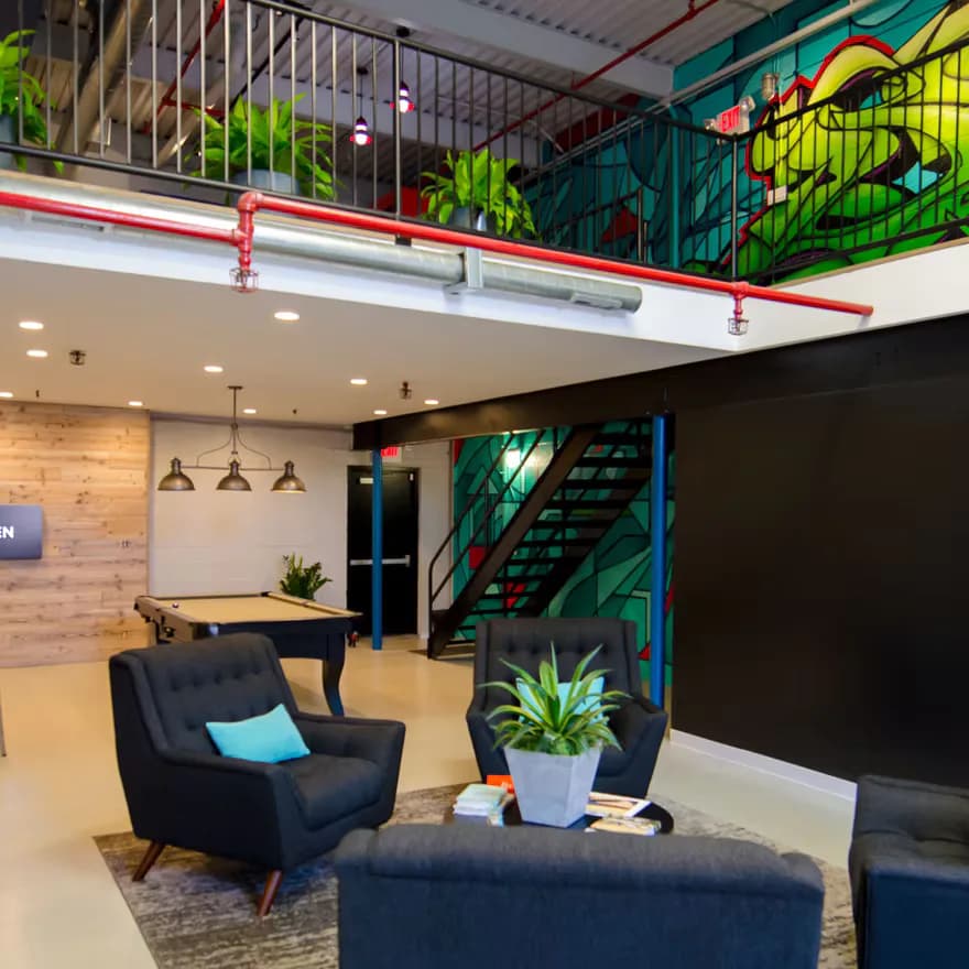 Modern office lobby with black accent wall, navy furniture, natural wood paneling, mezzanine level with green plants and red railings, designed by Mariama Agi.