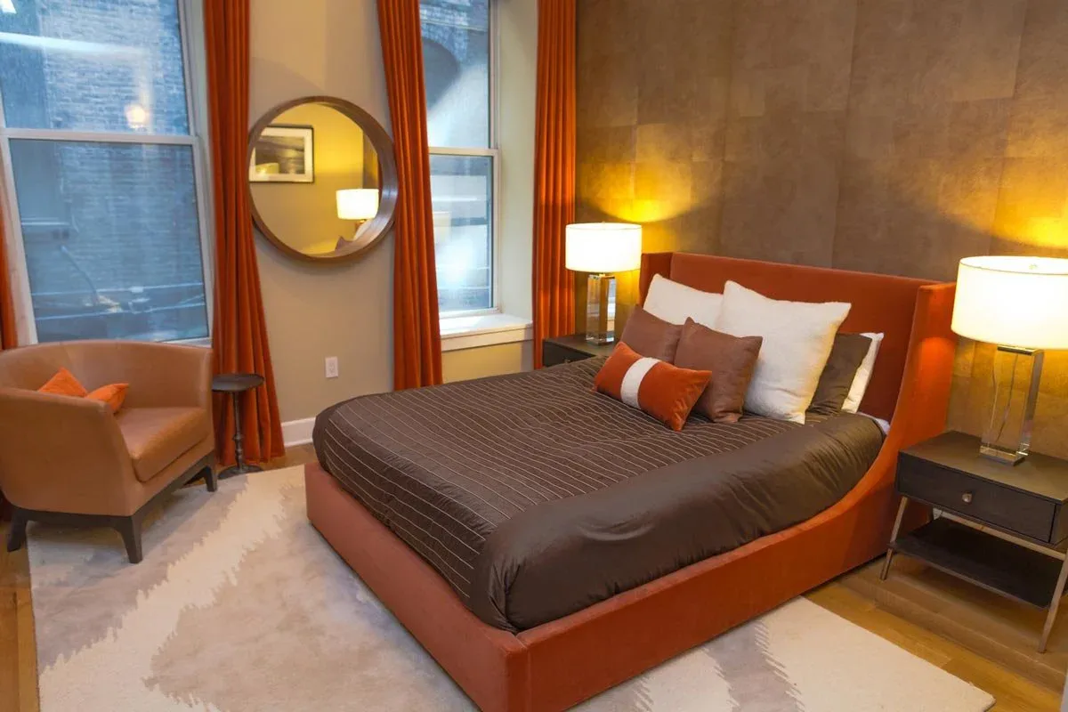Modern bedroom with orange upholstered bed frame, dark bedding, warm ambient lighting, round mirror, orange curtains, and mid-century furniture by Marie Burgos.