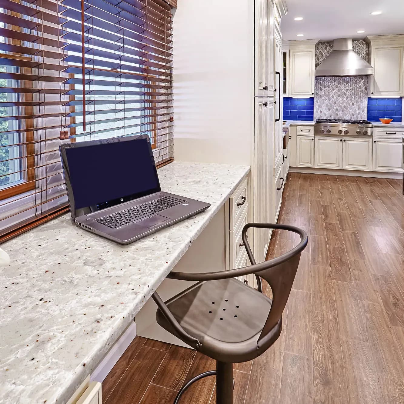 Modern kitchen workspace featuring a marble countertop with laptop and ergonomic chair, white cabinetry, blue backsplash accents, and wood flooring by Marilyn Ajose.