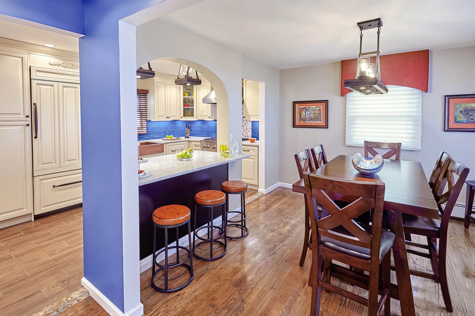 Open-concept kitchen and dining area with bold blue accent wall, wood dining table, breakfast bar with orange stools, and white cabinetry by Marilyn Ajose.