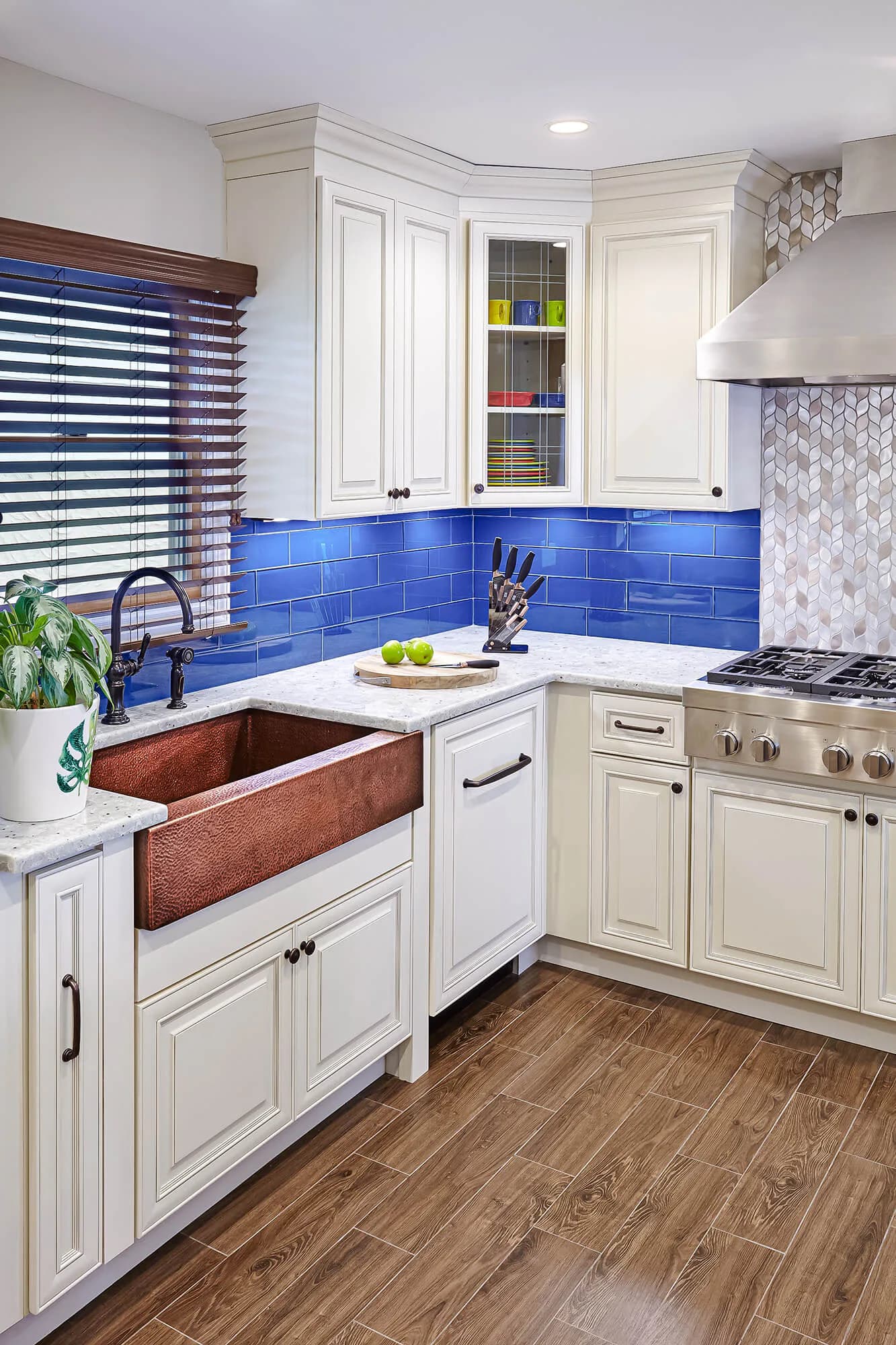 Contemporary kitchen with cream cabinetry, vibrant blue subway tile backsplash, copper farmhouse sink, and white countertops by Marilyn Ajose.