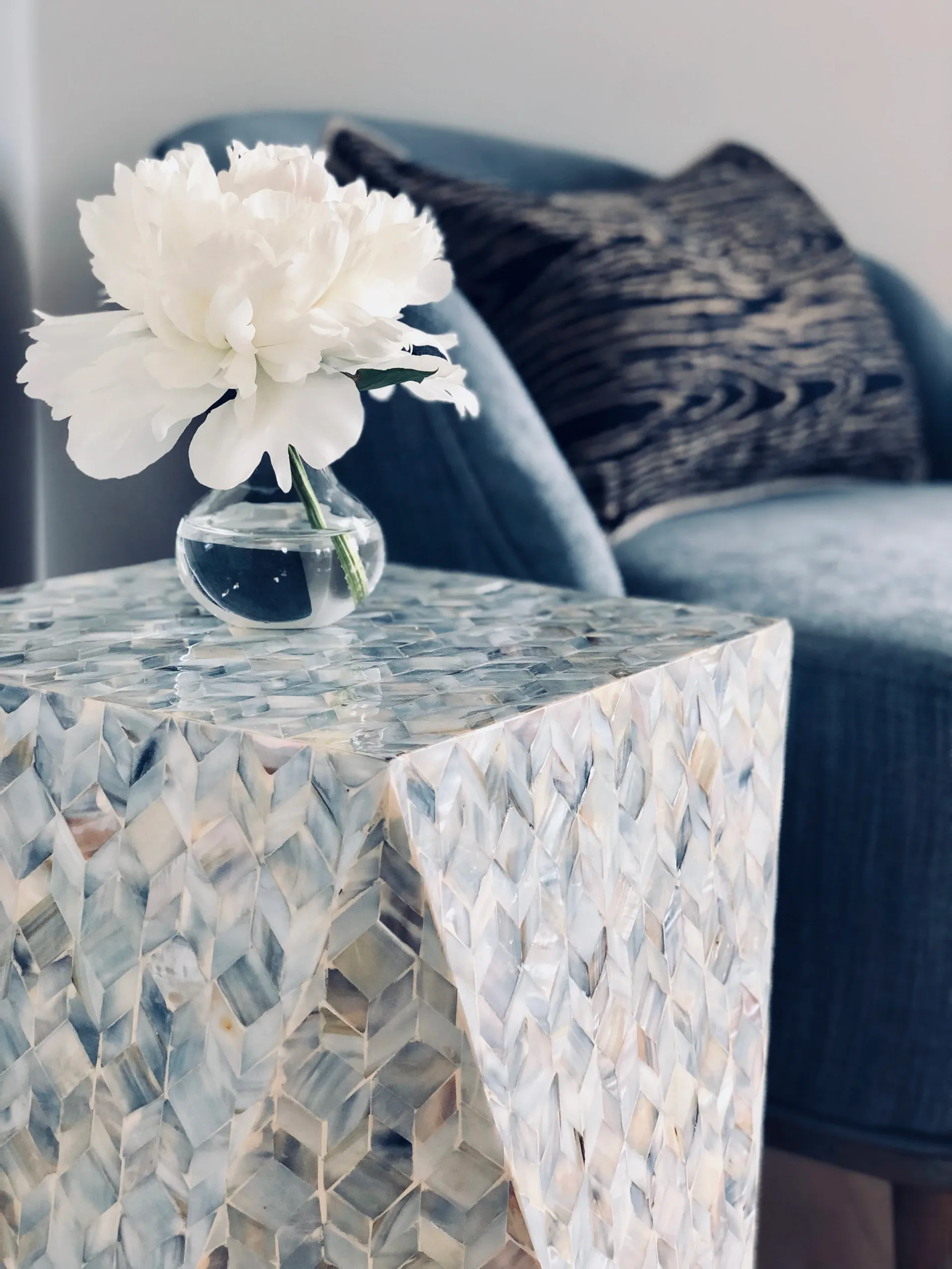 Minimalist side table with shell-pattern base holds a clear glass vase with white peony, styled with blue upholstered chair in neutral interior by Marina Mitchell.