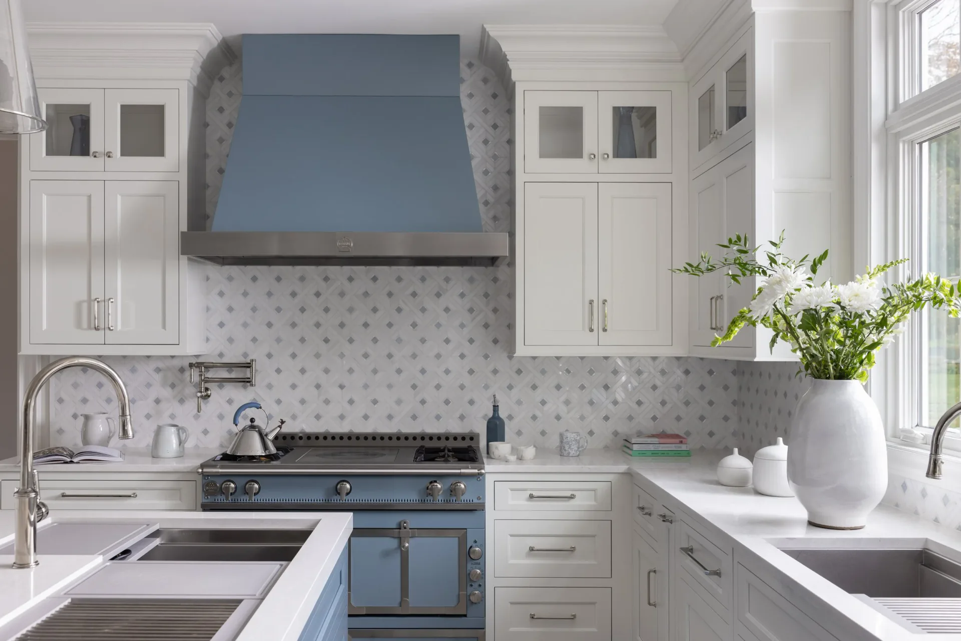 Bright kitchen with white cabinetry, blue range hood and stove, white marble countertops, and patterned backsplash by Marina Mitchell.