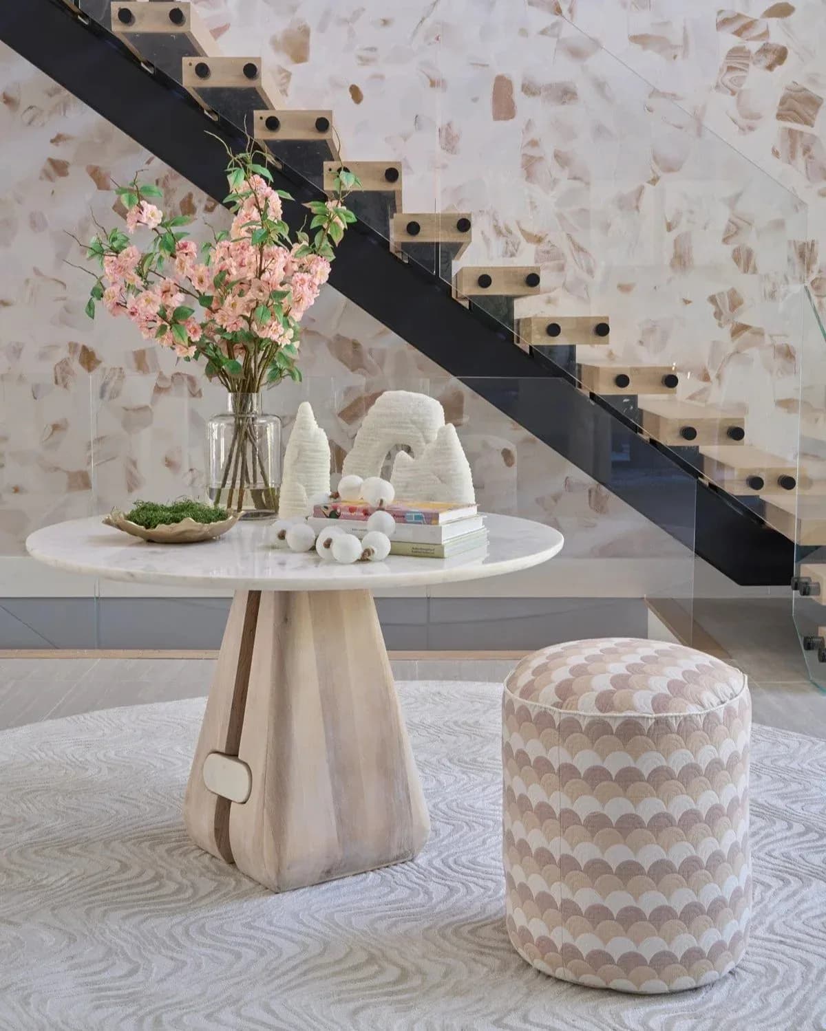 Modern entryway with terrazzo flooring, cream pedestal table with pink flowers, patterned ottoman, and black steel staircase against stone wall by Marina Mitchell.