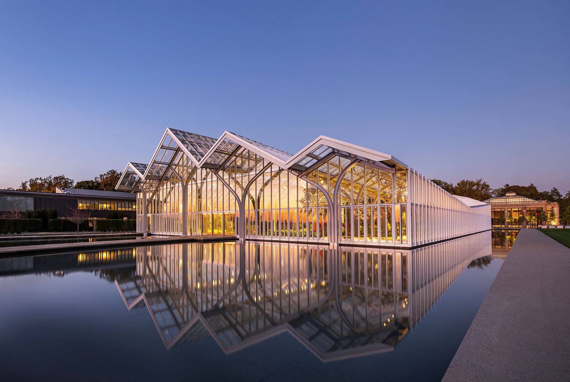 Modernist glass pavilion with white steel framework reflected in still water, illuminated at dusk, designed by Marion Weiss.