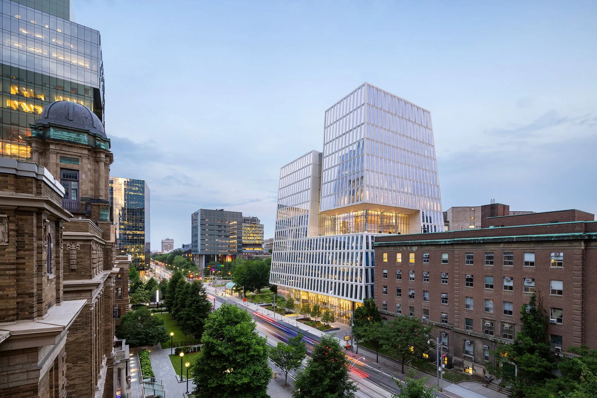 Urban streetscape at dusk showing modern mixed-use development with glazed towers and restored brick buildings along a tree-lined avenue by Marion Weiss.