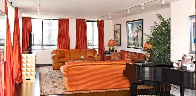 Vibrant living room with orange upholstered furniture, red curtains, black piano, patterned rug, and artwork by Marjorie Hilton.